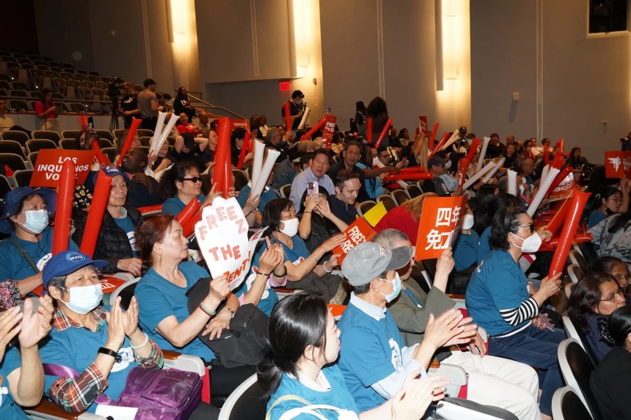 «Велика група людей у залі засідань тримає плакати та червоні повітряні трубки, закликаючи до заморозки орендної плати. Дехто тримає таблички з написами “FREEZE THE RENT” та гаслами різними мовами. Більшість учасників у синіх футболках, частина людей у масках. Атмосфера активістського зібрання, всі сидять у глядацьких кріслах і аплодують.»