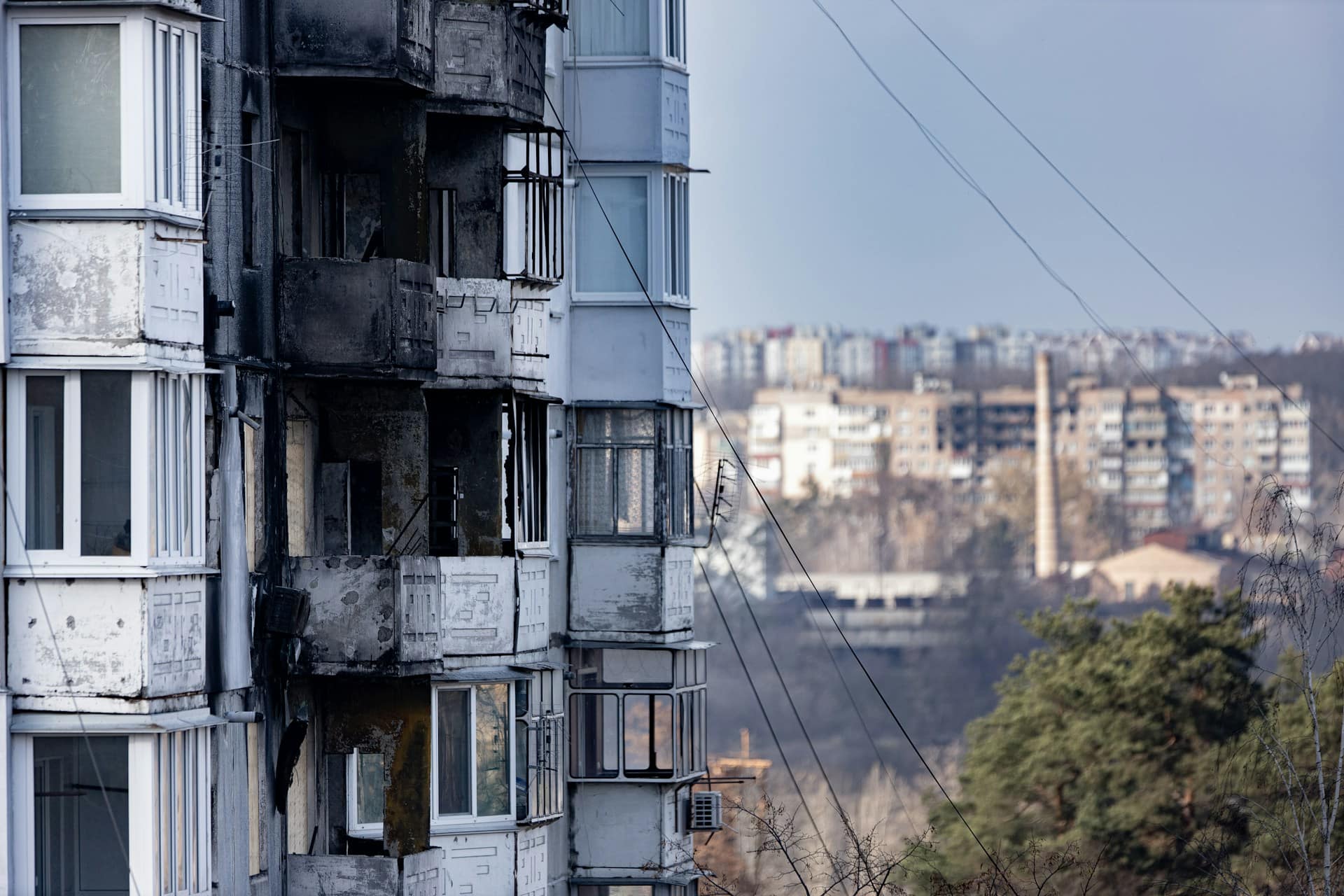Burned and damaged facade of a multi-storey apartment building with blackened balconies and windows, with a residential cityscape visible in the background.