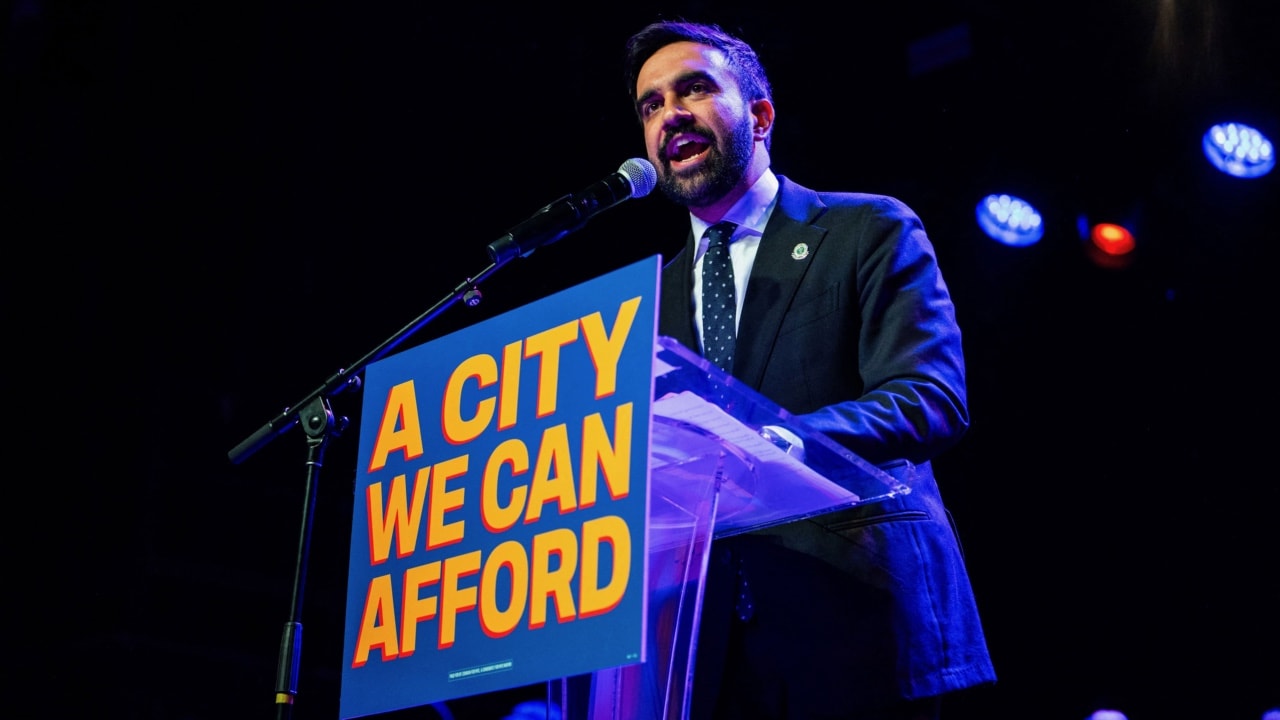 «Зохран Мамдані виступає на сцені за трибуною з мікрофоном. Перед ним стоїть синьо-оранжевий плакат із написом “A CITY WE CAN AFFORD”. Політик говорить у зал, позаду видно сценічні ліхтарі синього та червоного кольору.»